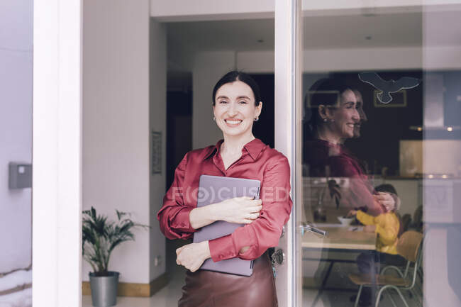 Femme d'affaires souriante tenant un ordinateur portable tout en se tenant à la porte en verre — Photo de stock