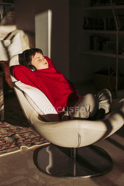 Niño escuchando música a través de auriculares mientras se relaja en la silla en casa - foto de stock