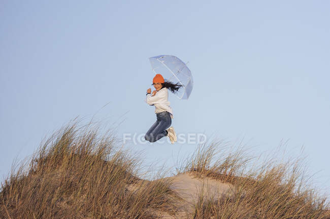 Молода жінка з парасолькою стрибає на вершині піщаної дюни проти неба — стокове фото