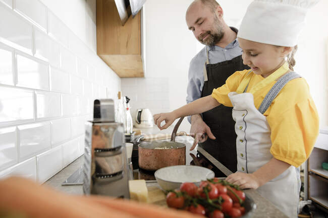 Figlia sorridente che indossa grembiule e cappello da chef che cucina il cibo mentre sta accanto al padre in cucina — Foto stock