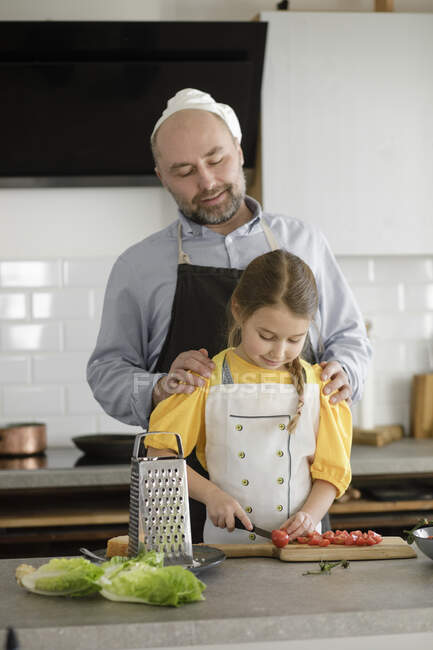 Padre mirando a su hija cortando tomate mientras está de pie en la cocina en casa - foto de stock