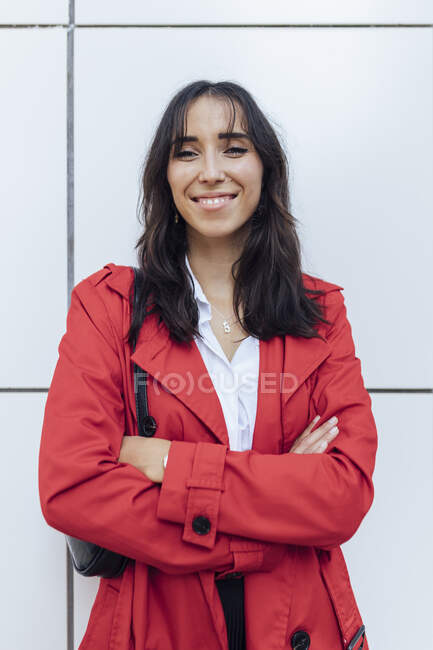 Mujer joven sonriente con gabardina roja de pie con los brazos cruzados contra la pared blanca - foto de stock