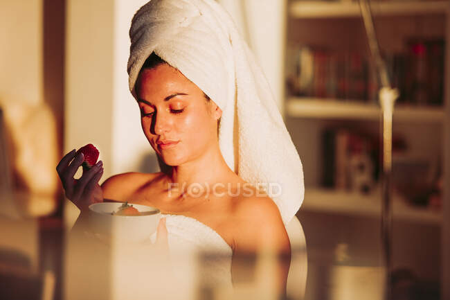 Woman in towel with fruit bowl having strawberry at home — Stock Photo