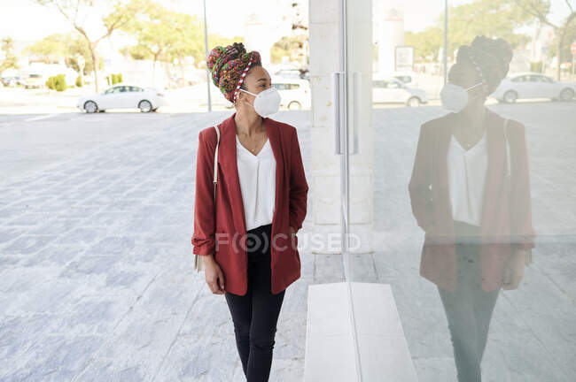 Mujer con pañuelo en la cabeza y mascarilla protectora mirando a la ventana mientras camina por la tienda - foto de stock