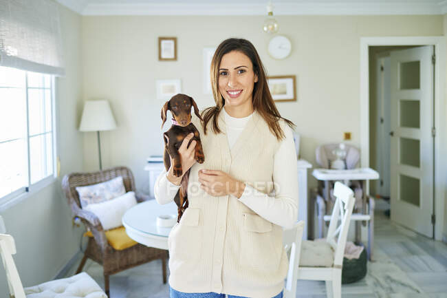 Smiling woman holding dog while standing in living room at home — Stock Photo