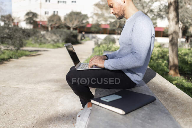 Homme d'affaires mature utilisant un ordinateur portable tout en étant assis sur un banc en béton parc public — Photo de stock
