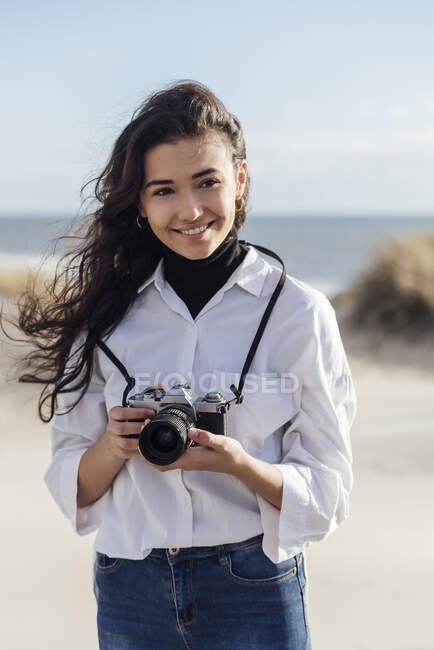 Посмішка красива жінка з вінтажною камерою стоїть на пляжі проти неба. — стокове фото