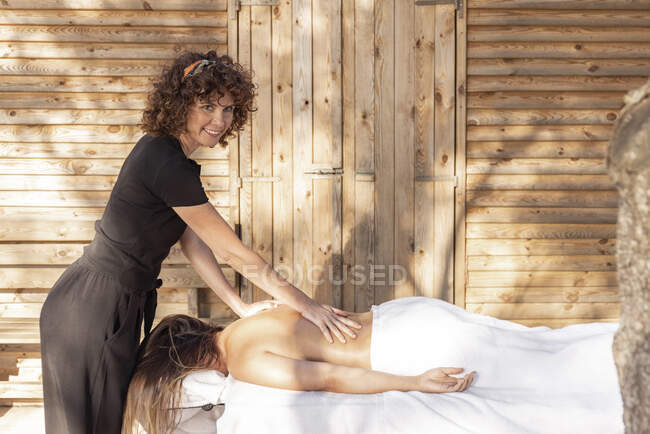 Massagista sorridente aplicando óleo sobre a mulher em pé ao lado da cabana — Fotografia de Stock
