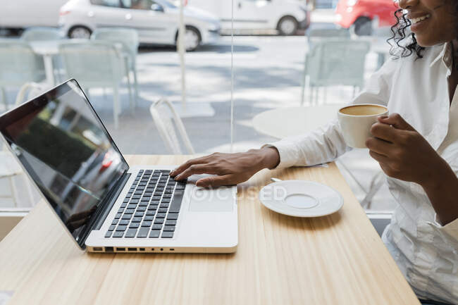 Жіночий фрилансер, який тримає чашку кави, користуючись ноутбуком у кафе. — стокове фото