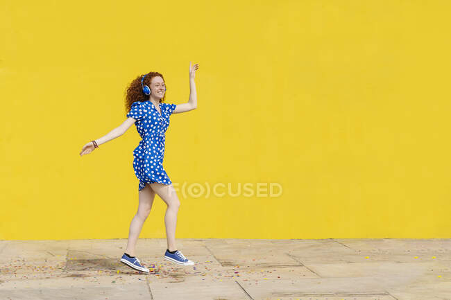 Carefree woman listening music while dancing by yellow wall — Stock Photo