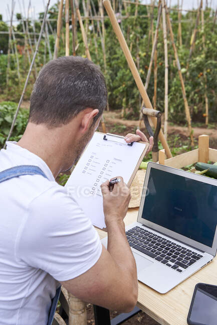 Male farmer writing on clipboard while sitting with laptop at table in agricultural field — Stock Photo