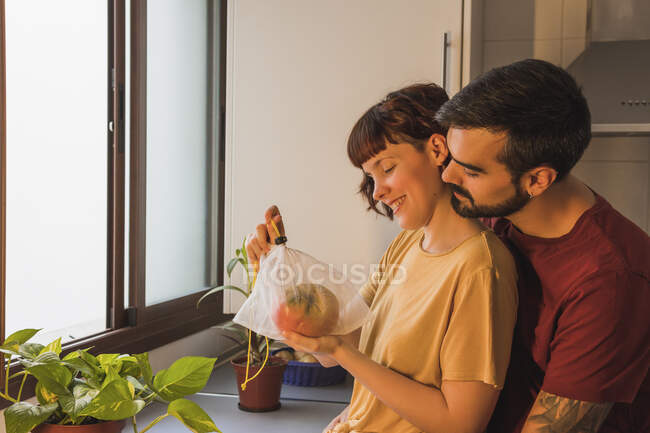 Man embracing girlfriend holding beefsteak tomato in kitchen at home — Stock Photo