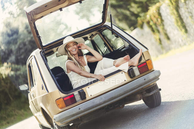 Happy blond woman wearing hat sitting in car trunk — Stock Photo
