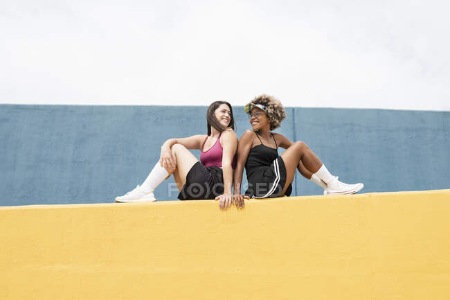 Souriant amis féminins se regardant tout en étant assis sur le mur de soutènement jaune — Photo de stock