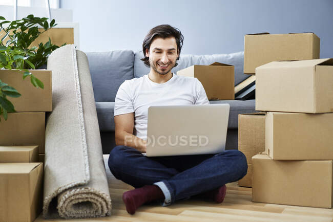 Uomo sorridente utilizzando il computer portatile mentre seduto in mezzo a scatole di cartone a casa — Foto stock
