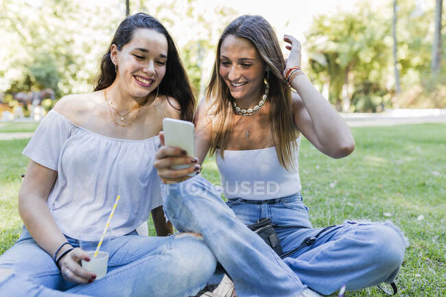 Smiling woman sitting by female friend sharing smart phone at park — Stock Photo