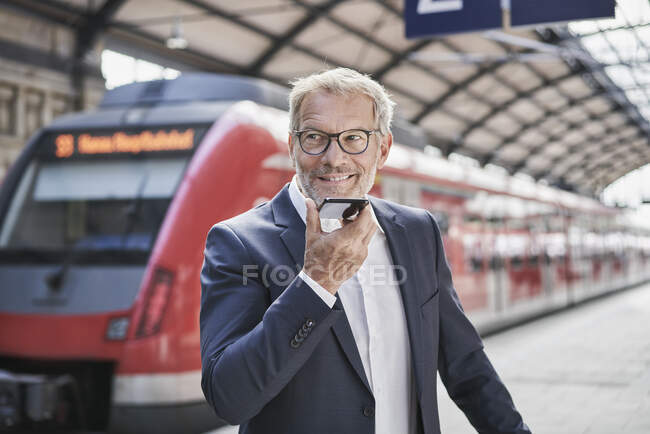 Smiling male professional looking away while sending voicemail through smart phone on railroad station — Stock Photo