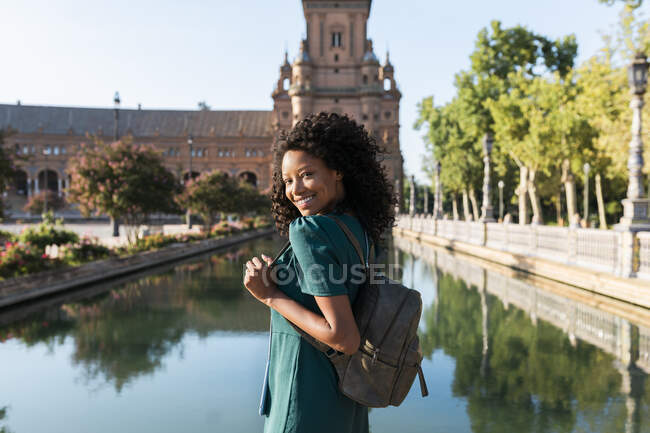 Giovane donna afro sorridente che indossa lo zaino vicino al laghetto a Plaza De Espana, Siviglia, Spagna — Foto stock