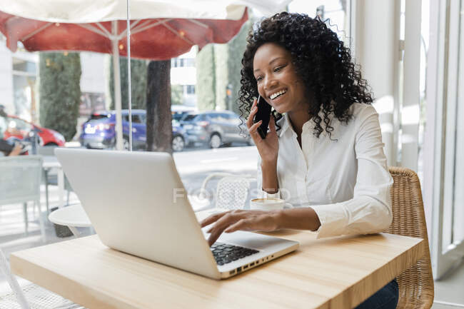 Бізнес-леді розмовляє на мобільному телефоні, використовуючи ноутбук у кав'ярні — стокове фото