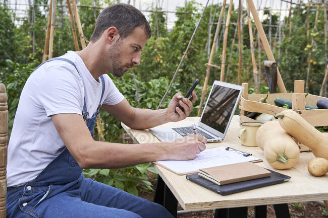 Male farm worker looking at smart phone while writing on clipboard sitting at table — Stock Photo