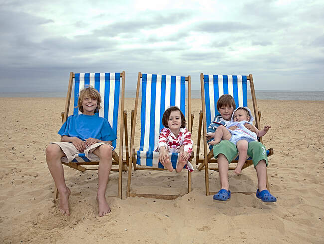 Brothers and sisters sitting on striped deck chairs at sandy beach during vacation — Stock Photo
