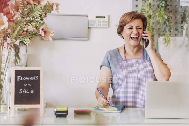 Cheerful female flower shop owner talking on mobile phone while standing at checkout — Stock Photo