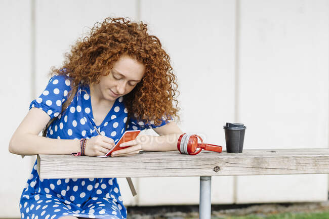 Mujer de pelo rizado escribiendo en bloc de notas mientras está sentada en la mesa - foto de stock