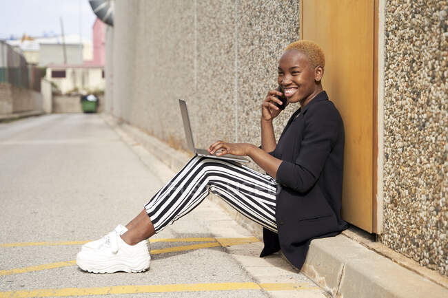 Sonriente profesional femenina con portátil trabajando en el teléfono a la luz del sol - foto de stock