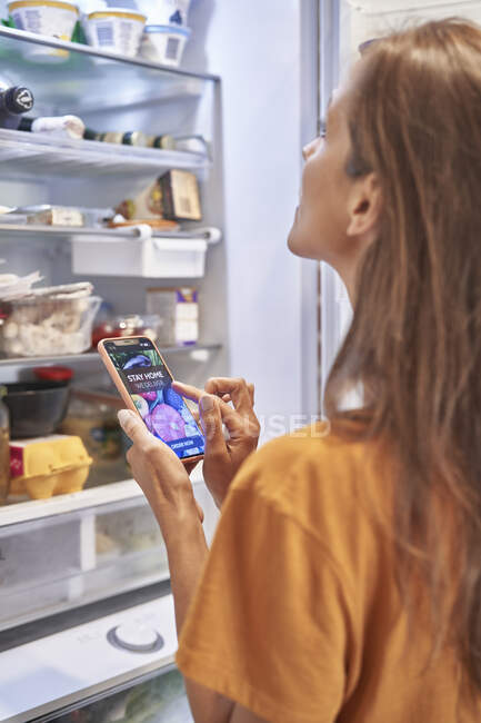 Donna che controlla il frigorifero mentre ordina generi alimentari attraverso l'applicazione mobile — Foto stock