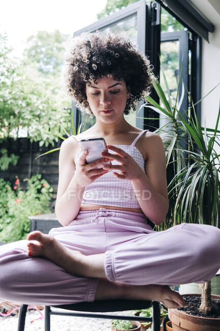 Молодая женщина с помощью мобильного телефона, сидя скрестив ноги на стуле — стоковое фото