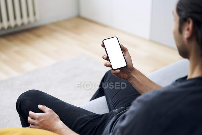 Мужчина держит смартфон, сидя дома на диване — стоковое фото