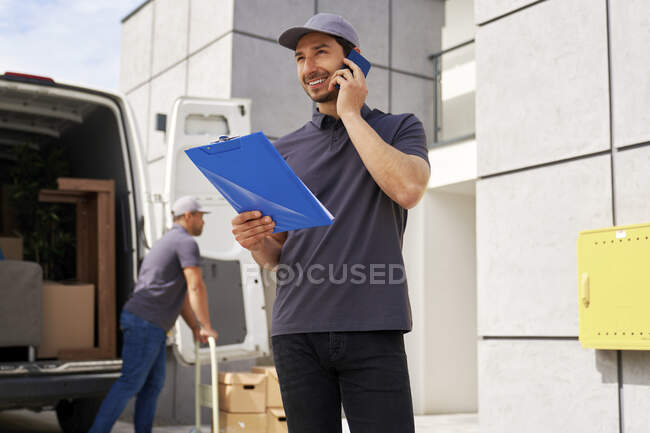 Souriant livreur tenant presse-papiers tout en parlant sur téléphone intelligent — Photo de stock
