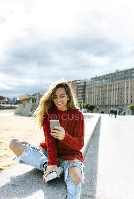 Femme heureuse utilisant un téléphone intelligent tout en étant assis sur le mur de soutènement — Photo de stock