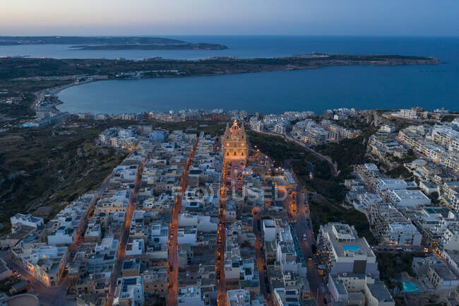 Malta, Nördliche Region, Mellieha, Luftaufnahme der Küstenstadt in der Abenddämmerung mit beleuchteter Pfarrkirche Mariä Geburt im Hintergrund — Stockfoto