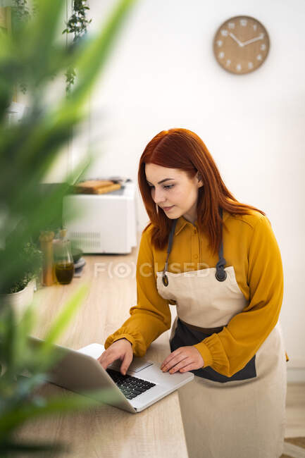 Redhead woman with apron using laptop while standing in kitchen at home — Stock Photo