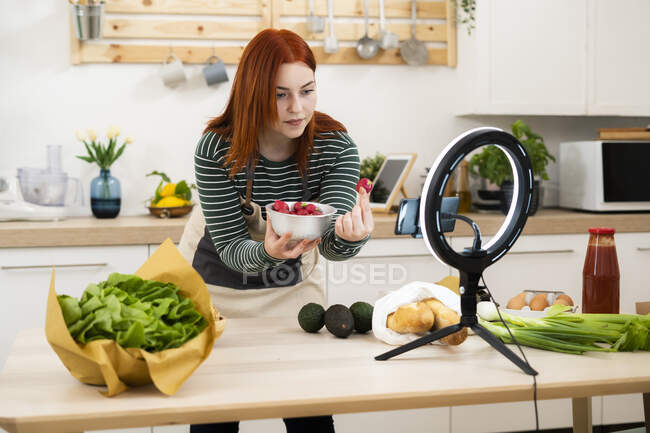 Woman showing strawberry while live streaming through mobile phone in kitchen — Stock Photo