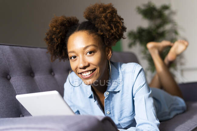 Усміхнена жінка Афро з цифровим планшетом лежить на дивані у вітальні — стокове фото