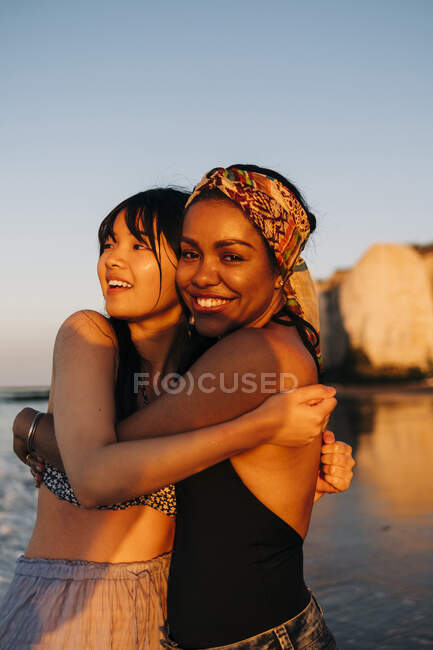 Sonrientes amigas abrazándose en la playa - foto de stock