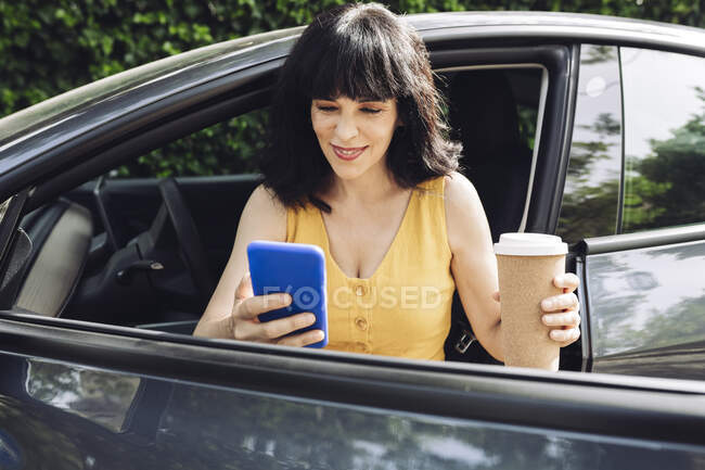 Mujer feliz con taza de café desechable usando el teléfono inteligente mientras sale del coche eléctrico - foto de stock