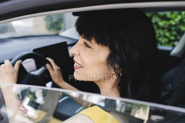 Mujer madura sonriente mirando desde la ventana mientras conduce un coche eléctrico - foto de stock