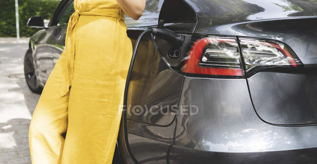 Mujer madura apoyada en coche eléctrico en la estación de carga - foto de stock