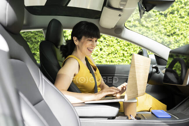 Happy businesswoman using laptop while sitting in electric car — Stock Photo