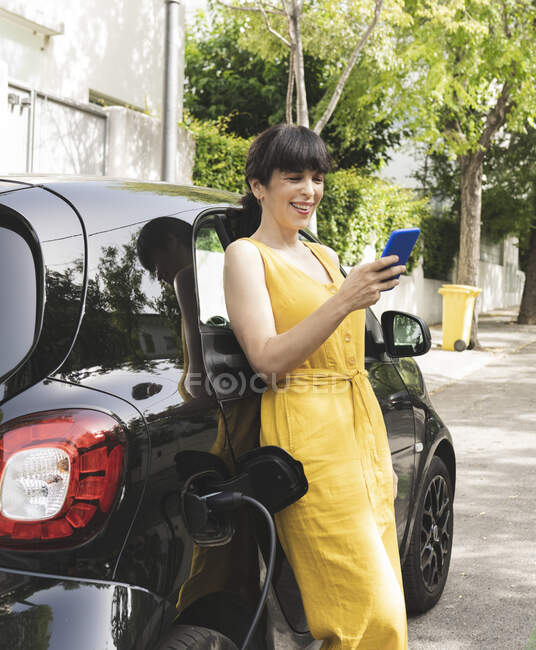 Smiling mature woman using smart phone at electric car charging station — Stock Photo