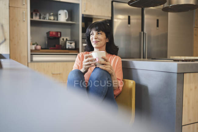 Улыбающаяся зрелая женщина с чашкой кофе сидит на кухне дома — стоковое фото