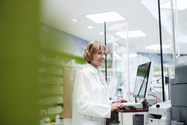 Farmacêutica feminina sorridente em pé à mesa na farmácia — Fotografia de Stock