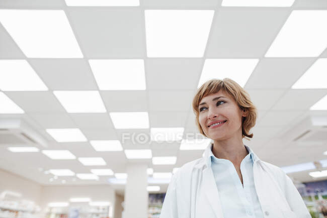 Farmacéutica sonriente mirando hacia otro lado mientras está parada en la farmacia - foto de stock