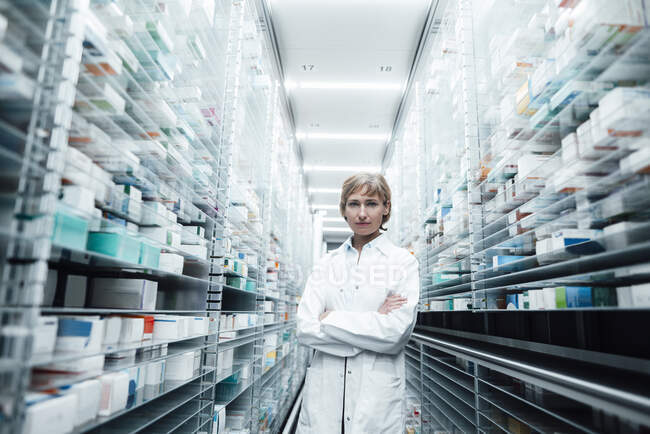 Fiduciosa farmacista donna con le braccia incrociate in piedi in mezzo a scaffali in negozio medico — Foto stock