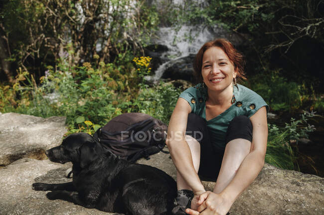Sorrindo médio adulto proprietário do sexo feminino sentado ao lado do cão na floresta — Fotografia de Stock