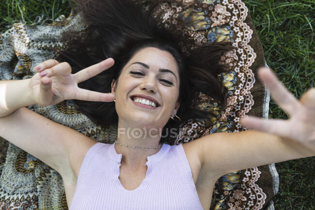 Happy young woman gesturing peace sign while lying at public park — Stock Photo