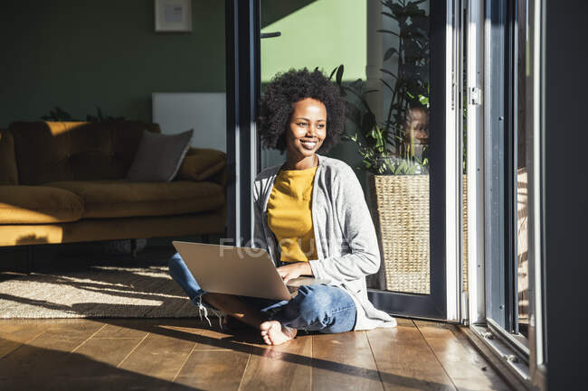 Lächelnde junge Frau mit Laptop sitzt zu Hause im Sonnenlicht — Stockfoto